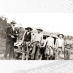Seis homens posam diante de um automóvel antigo, em fotografia histórica em preto e branco.