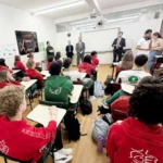 Alunos sentados em carteiras numa sala de aula, com profissionais em pé ao fundo.