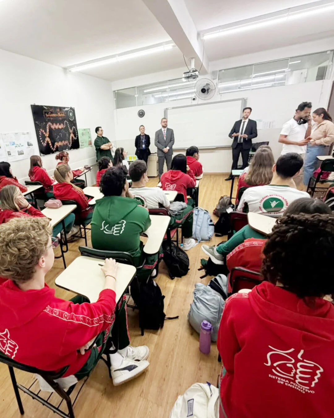 Alunos sentados em carteiras numa sala de aula, com profissionais em pé ao fundo.