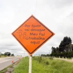 Placa laranja à beira da rodovia com a frase "Por favor vai devagar. Meu pai trabalha aqui!".