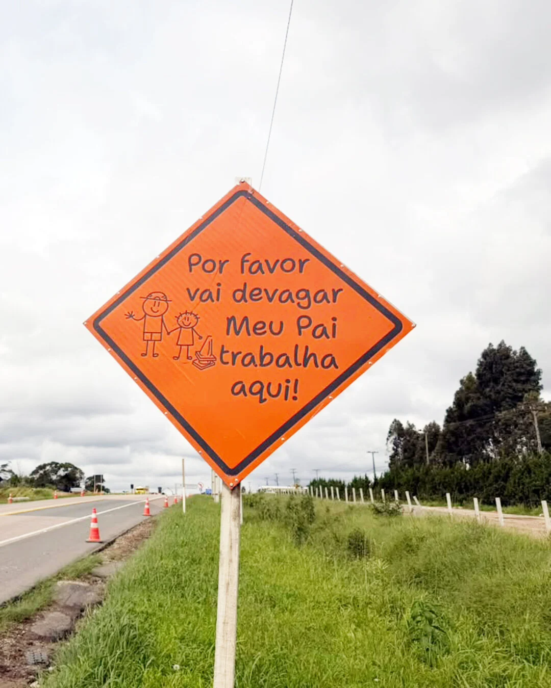 Placa laranja à beira da rodovia com a frase "Por favor vai devagar. Meu pai trabalha aqui!".